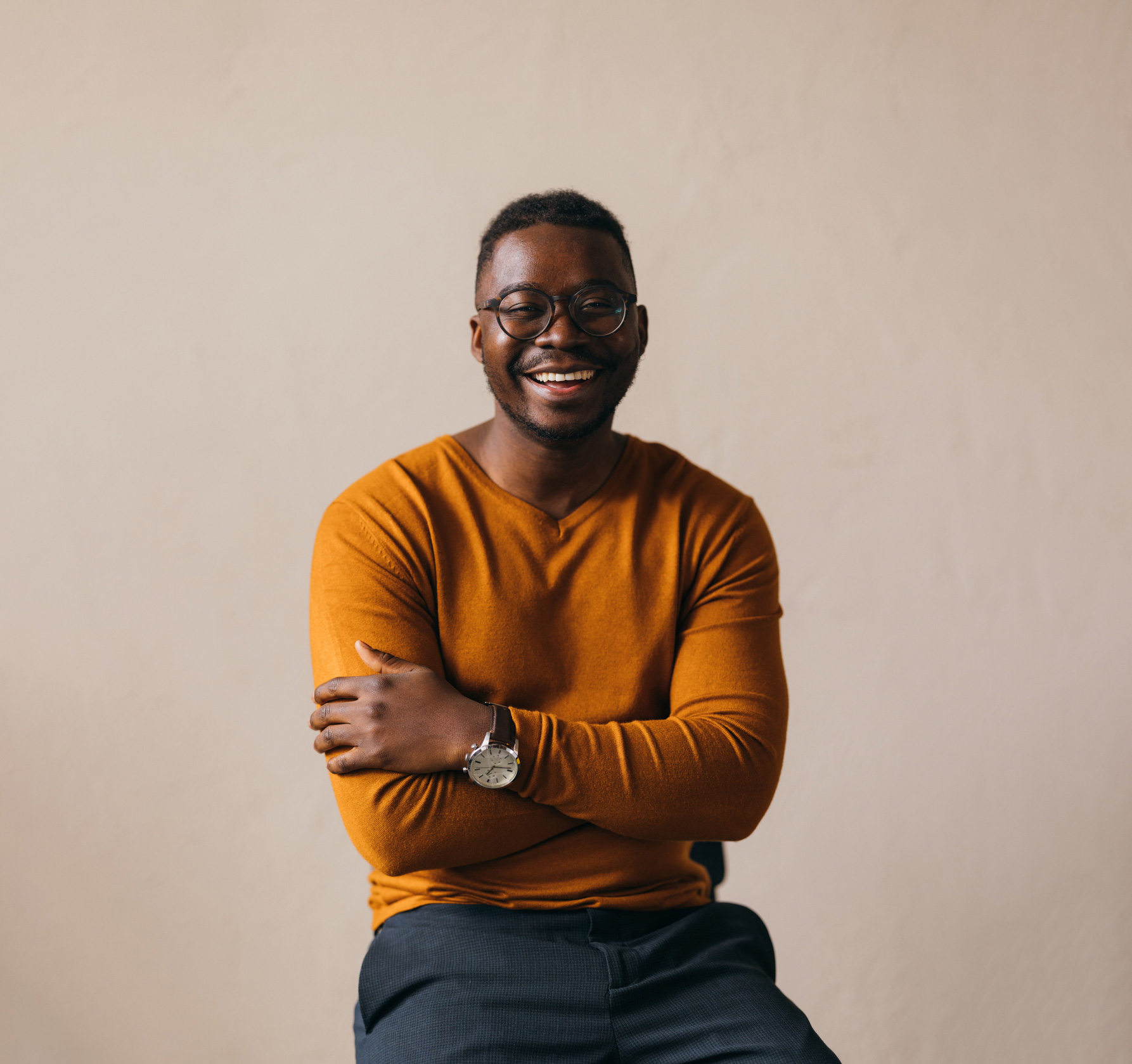 A smiling man with glasses, seated, with an orange sweater and charcoal trousers.